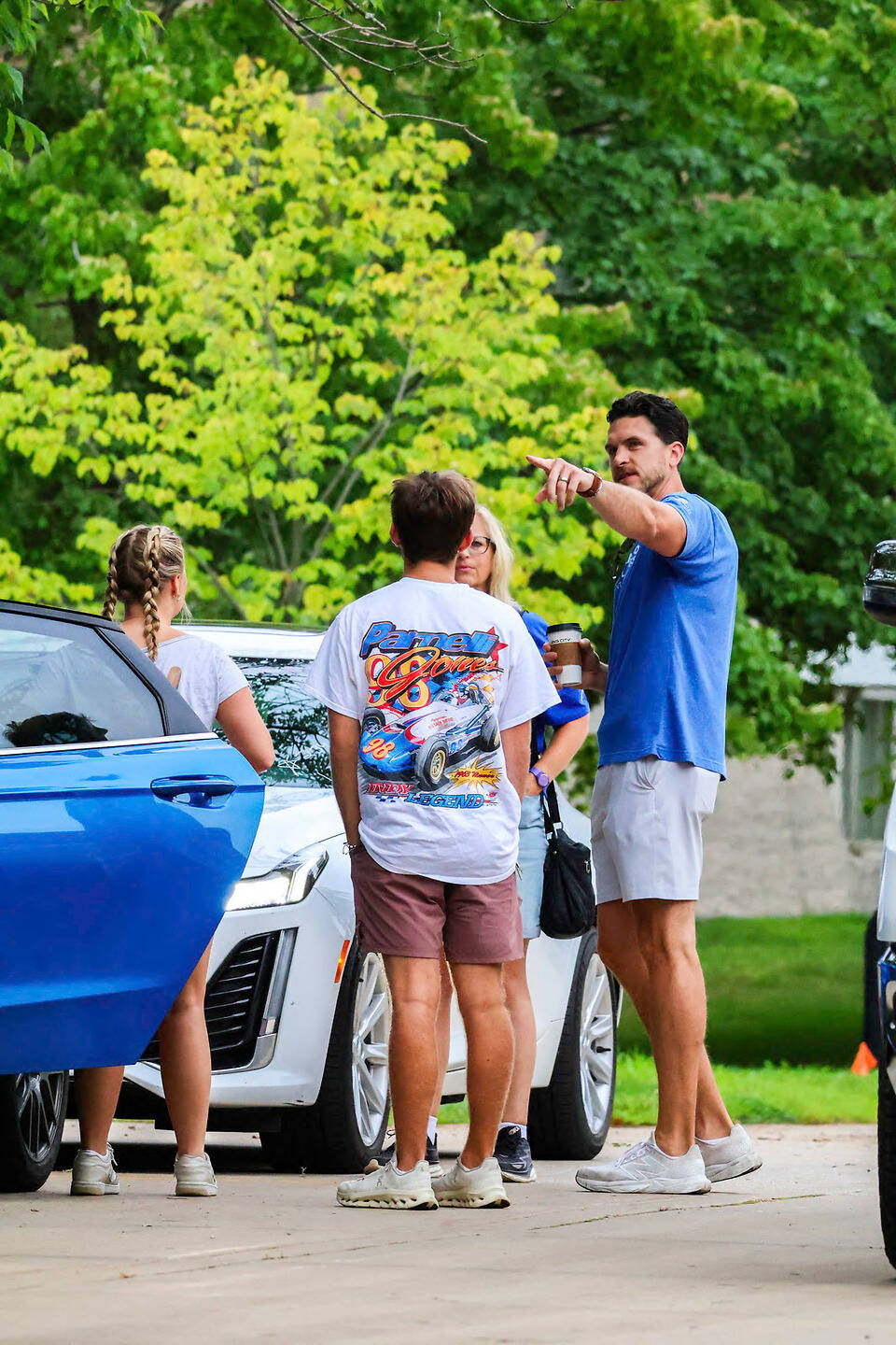 A group of people stand near parked cars in a driveway, with one person pointing while holding a coffee cup.